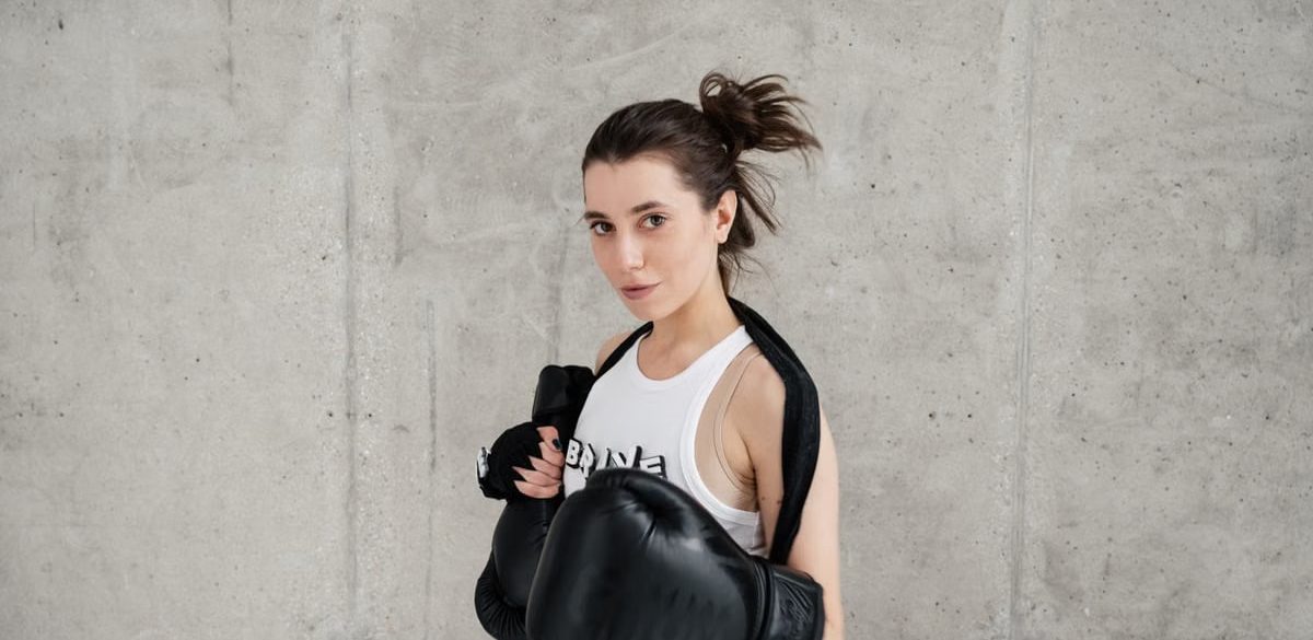 Confident young woman posing with boxing gloves against a concrete wall, showcasing style and sportiness.