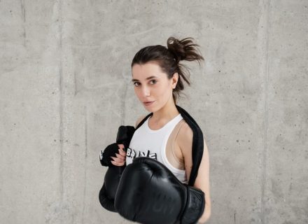 Confident young woman posing with boxing gloves against a concrete wall, showcasing style and sportiness.