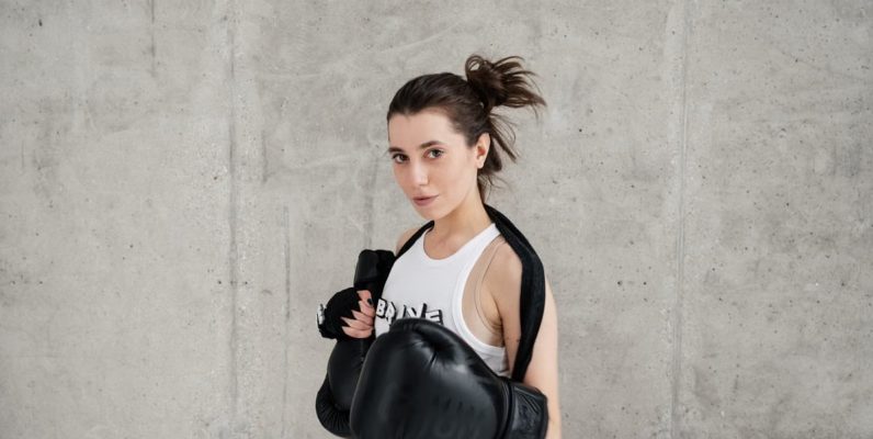 Confident young woman posing with boxing gloves against a concrete wall, showcasing style and sportiness.